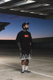 Man wearing a black t-shirt with red text and shorts in an open area with a blue sky.